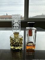Two vintage perfume bottles on table by window, one square cut glass with etched floral stopper, the other rectangular with black lid and red label