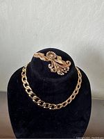 Photo showing gold-tone chain necklace with large oval links and vintage-style brooch with pearl accents on black display.