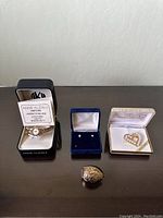 Full view of the six jewelry items on a dark surface, including watch in box, earrings in blue velvet box, brooch in Jennifer Moore box, ring in front