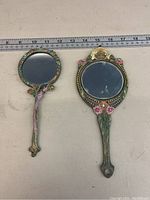 Front view of two decorative hand mirrors with floral and bead embellishments placed next to a measuring tape showing lengths around 7-8 inches.