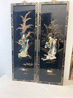 Two vertical wood panels with black backgrounds featuring stone-carved ladies in colorful traditional clothing and gold leaf botanical designs.