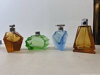 Four art deco crystal perfume bottles in amber, green, blue, and amber displayed in a row, showing geometric faceted shapes and metal pump caps but no bulbs.