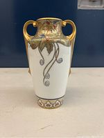 Front view of a white arts and crafts style porcelain vase with gold handles and floral lemon leaf patterns.