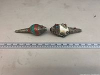 Top view of two decorative conch shells with detailed silver-tone metalwork, colorful stone inlays and measuring tape showing length.