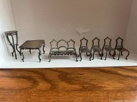 Seven sterling silver miniature dollhouse furniture pieces displayed in a row on a white background: one tall round pedestal table, one rectangular table, one loveseat sofa, and four chairs with ornate backs.