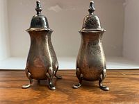 Front view of the two sterling silver shakers standing side by side, showing shape, legs, and tops.