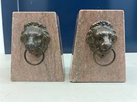 Pair of trapezoid stone bookends with mounted lion head metal rings, visible chip on corner of one bookend.