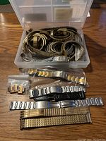 Open plastic box filled with metal stretch expandable watch bands in mixed silver and gold tones. Below the box are various metal link bands in silver and gold tones, two black leather watch straps, and smaller plastic bags containing spare links and clasps.