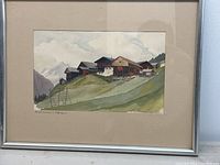 Framed watercolor painting depicting a mountain town scene with traditional buildings on a hillside and mountains in the background.