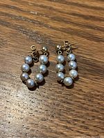 Close-up photo of a pair of 14kt gold earrings with white pearls arranged along each hoop, laying on a wooden surface.