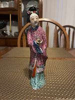 Front view of porcelain Geisha figurine showing detailed face, hair ornaments, and pink kimono with blue sleeves.