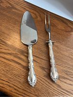 Photo showing a silver-toned cake/pie server and a two-prong serving fork both with ornate sterling silver handles, placed on a wooden surface.