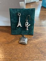 Photo shows a square sterling silver locket with engraved detail lying flat and two sterling silver earrings on a dark green display stand. One earring is shaped like the Eiffel Tower and the other like a figurine.