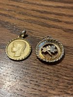 Two pendants on wood surface; gold-tone pendant with profile and round two-tone pendant with floral design.