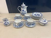 Photo showing entire coffee set including 6 side plates stacked, 4 tea cups on saucers, sugar bowl with lid, creamer jug, and urn with lid, all with blue onion pattern on white porcelain.