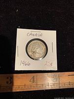 Obverse side of 1966 Canada 25 cents coin showing Queen Elizabeth II and inscriptions, coin secured in cardboard holder with visible markings 'CANADA', '1966' and '25¢'. A ruler placed nearby shows the coin is about 23 mm in diameter.