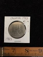 Obverse side of the 1983 Canada dollar coin with Queen Elizabeth II profile and date '1983'. Coin in a white cardboard holder with handwritten text.