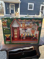 Front boxed photo of the Northpole Station animated Christmas decoration showing the dollhouse-style building with clock tower, Santa and elf figures behind window, and the 'Northpole Station' branding on the box.