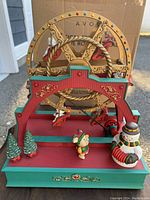 Vintage Avon musical Christmas Ferris wheel figurine with snowman and bear, lights and decorated evergreen trees, on a wooden base with decorative gold trim, original box in background.