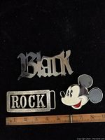 Front view of three metal belt buckles showing 'Black' text, 'ROCK' text, and Mickey Mouse head enamel buckle. Ruler for scale included