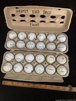 Two dozen white golf balls arranged in two cardboard holders, 12 per holder, showing brands and general condition