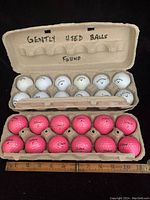 Two cartons each holding a dozen golf balls; top carton contains 12 white Callaway golf balls, bottom carton contains 12 pink Titleist golf balls.
