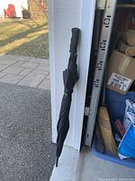 Closed black Nautica golf umbrella leaning against a garage entrance near a broom and storage bins.