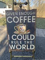 Close-up image of the wooden coffee wall art sign held on a brass easel outdoors, showing visible text and coffee cup image.