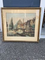 Framed etching of a European village scene with a stone bridge and buildings along a canal, shown outside against a paved surface.