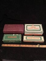 Top view of four boxed Singer sewing machine kits including a red plastic case, two green and white boxes, and a stitch pattern set box with a measuring ruler for scale.
