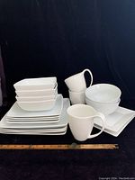Full set of stacked square plates and bowls with mugs placed in front; shows quantity and style of dinnerware