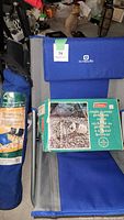 Photo of two camping chairs, one OUTWELL brand in blue and grey, and an ESCORTE Deluxe Sport Chair in blue, both with carrying bags. Coleman single burner stove in green box placed on one chair.