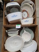 Two cardboard boxes containing CorningWare Blue Cornflower casseroles with lids along with mixed ceramic and Corelle dinnerware pieces