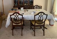 View of the mahogany dining table with two chairs showing the table covered with a protective layer and the carved wooden chairs with lattice pattern backs.