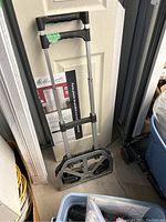 Foldable dolly shown in folded position leaning against wall, metal frame, plastic wheels, black cushioned handle visible.