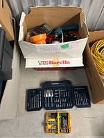 Box containing assorted tools and drill accessories, with two sets of drill bits cases on floor below.