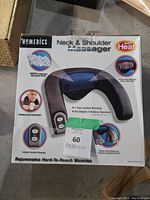Front view of the boxed HoMedics neck and shoulder massager showing the product image and features such as 2-speed massage, heat option, cushioning microbeads, and remote control buttons.