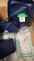 Box with multiple Pyrex glass rectangular food storage containers and navy blue lids. Some lids stacked and some containers nested inside others. Containers appear clean but seller notes they need cleaning.