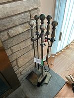 Six metal fireplace tools with ball-top handles placed upright in a metal stand on hearth near stone fireplace.
