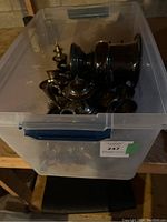 Plastic bin filled with assorted tarnished silver and silver-plated kitchenware items, including teapots, a large urn, cups, pitchers, and other vessels.