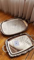 Two trays side by side on wooden floor, one silver plated with floral engraving, one brass with smooth surface.