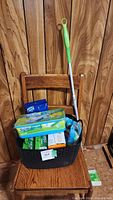 Full view of black plastic basket containing Swiffer mop, several boxes and packages of Swiffer cleaning pads, and cleaning sponges set on wooden chair.
