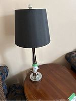 Table lamp with black shade on a silver base, shown on a wooden table beside furniture.