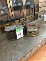 Two vintage charcoal irons side by side on stone surface in front of glass cabinet. Larger iron has black handle and decorative embossing, smaller iron has slotted body and wooden knob handle.