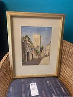 Full view of framed watercolor painting displayed against a blue wall and wicker chair.