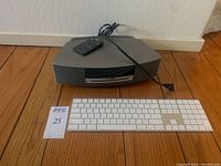 Gray Bose Wave Music System III stereo with remote and white Apple wireless keyboard on wooden floor.