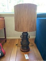 Full view of the lamp showing the carved metal base, wood base, woven lampshade, and electrical cord on wooden floor near furniture.