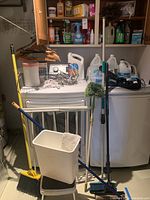 Photo of laundry and cleaning supplies on top of washer and dryer, showing mops, brooms, bucket, extension cords, Wahl haircutting tool, and cleaning liquids.