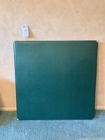 Full view of the vintage green card table folded flat and standing upright against a wall, showcasing the textured green surface.