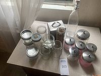 Collection of clear and pink glass jars with metal lids, a tall glass bottle with swing top clasp, a square metal box, and small bundle of wooden sticks on a windowsill.
